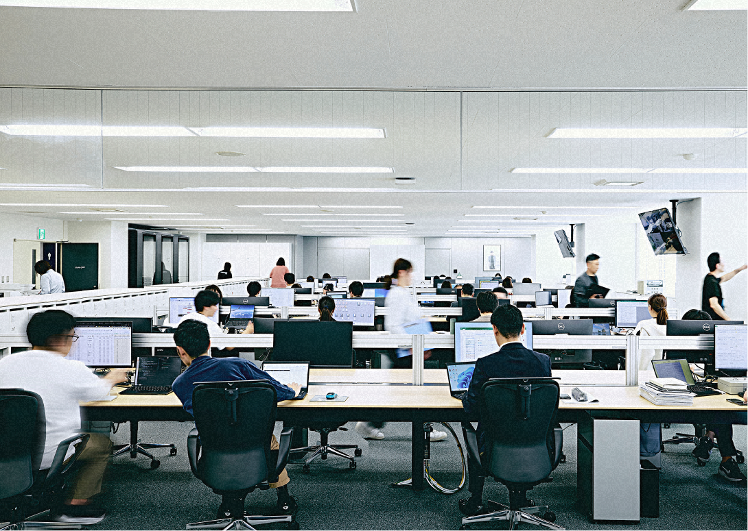 パソコンが並んだ大きなオフィスの様子。長机に複数のパソコンと椅子が整然と配置されている写真